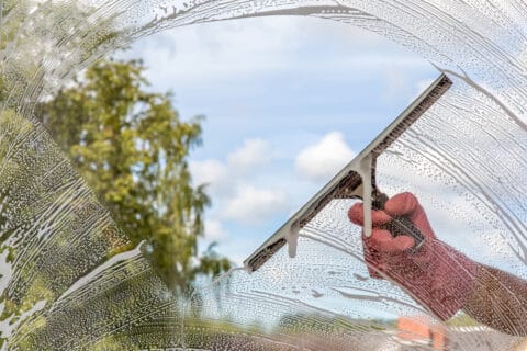 Hand in protective glove washing and cleaning window with professionally squeegee on background of cloudy sky. Summer windows cleaning. Maid cleans window.
